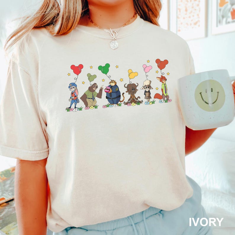 May include: Ivory-colored t-shirt featuring a whimsical cartoon design of several animal characters holding heart-shaped balloons. The characters are standing on a patch of grass with stars above them. A white mug with a smiley face is on the right.