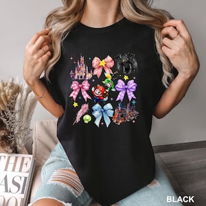 May include: Black crew neck t-shirt featuring a colorful graphic design. The design includes illustrations of castles, bows, and characters. The word "BLACK" is printed at the bottom right corner of the shirt.