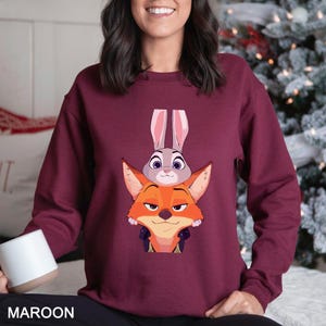 Pode incluir: Camisola bord&ocirc; com um desenho de desenho animado de uma raposa e um coelho. A raposa &eacute; laranja, o coelho &eacute; cinza. A palavra "MAROON" &eacute; impressa na parte inferior da camisola. Um design festivo e divertido.