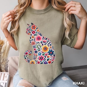 May include: Khaki-colored t-shirt featuring a floral cat silhouette. The cat design is filled with colorful flowers in shades of pink, yellow, blue, and red. The shirt has a crew neck and short sleeves. The word "KHAKI" is printed at the bottom.