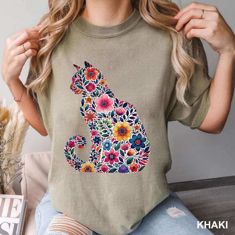 May include: Khaki-colored t-shirt featuring a floral cat silhouette. The cat design is filled with colorful flowers in shades of pink, yellow, blue, and red. The shirt has a crew neck and short sleeves. The word "KHAKI" is printed at the bottom.