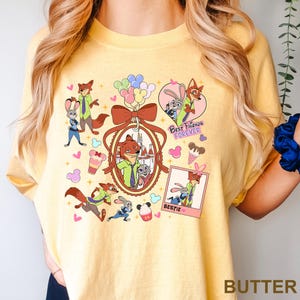 Op de afbeelding: Boterkleurig T-shirt met een kleurrijke afbeelding van cartoon vos- en konijnfiguren. Het ontwerp bevat de tekst "Best Friends Forever" en "Bestie", samen met ijsjes, ballonnen en harten. Het shirt heeft een ronde hals.
