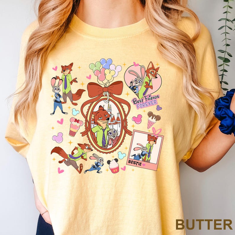 Op de afbeelding: Boterkleurig T-shirt met een kleurrijke afbeelding van cartoon vos- en konijnfiguren. Het ontwerp bevat de tekst "Best Friends Forever" en "Bestie", samen met ijsjes, ballonnen en harten. Het shirt heeft een ronde hals.