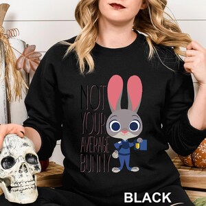 Op de afbeelding: Zwarte crewneck sweatshirt met een cartoon konijn karakter in een blauw uniform en roze oren. De tekst "NOT YOUR AVERAGE BUNNY" is gedrukt in zwart en roze. Het woord "BLACK" staat onderaan.