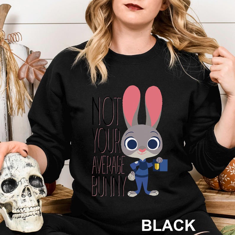 Op de afbeelding: Zwarte crewneck sweatshirt met een cartoon konijn karakter in een blauw uniform en roze oren. De tekst "NOT YOUR AVERAGE BUNNY" is gedrukt in zwart en roze. Het woord "BLACK" staat onderaan.