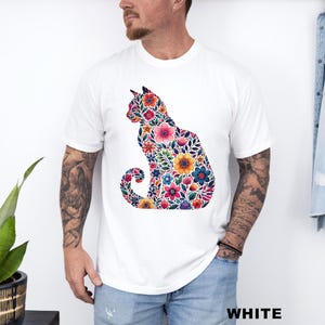 May include: White t-shirt featuring a colorful floral cat design. The cat silhouette is filled with various flowers in shades of pink, orange, blue, and yellow. The word "WHITE" is printed in bold, black letters at the bottom of the image.