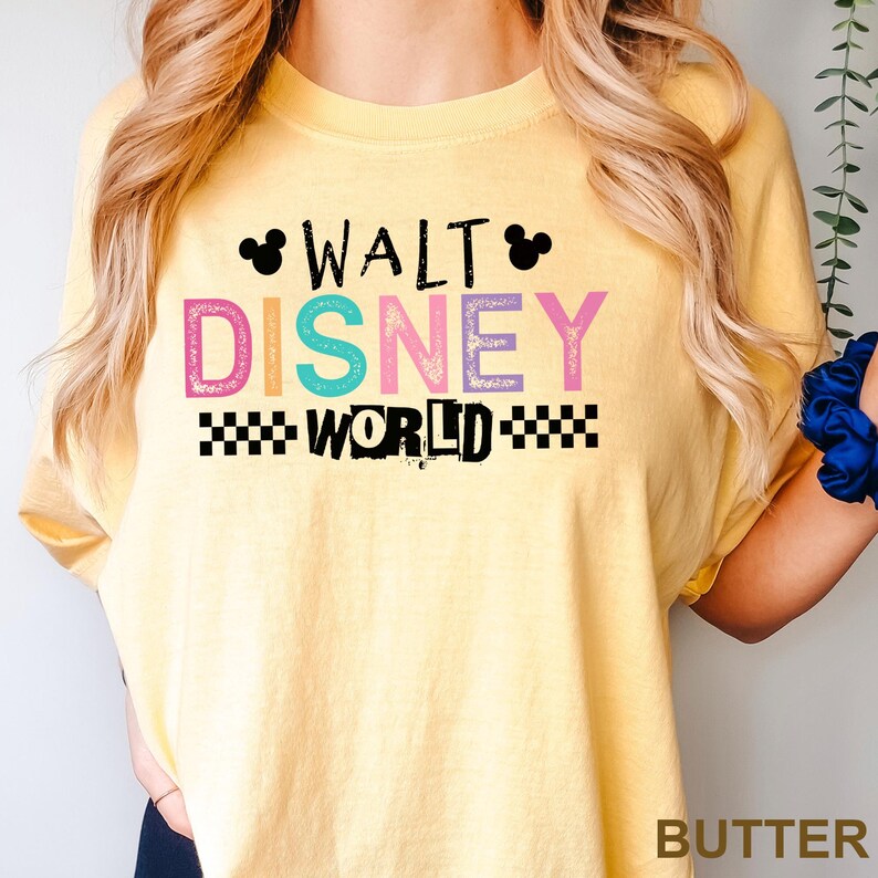 May include: A butter yellow t-shirt featuring the text WALT DISNEY WORLD in a mix of black and colorful, textured letters. The design includes two small black Mickey Mouse ear silhouettes above the word WALT and black checkered patterns.