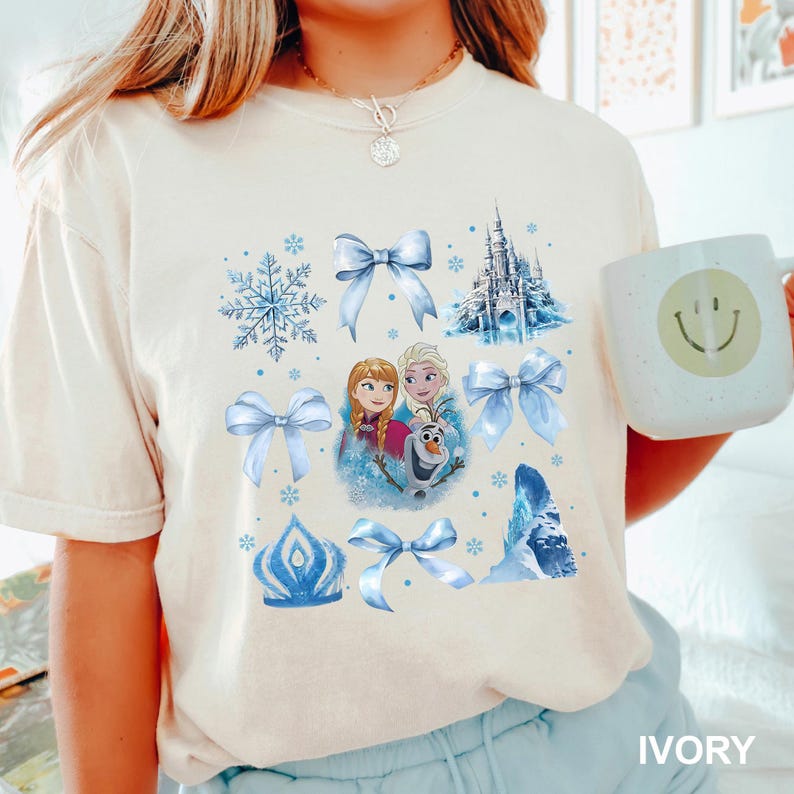 May include: Ivory-colored t-shirt featuring a collage of blue snowflake, bow, castle, and character graphics. The shirt includes a graphic of Anna, Elsa, and Olaf. A white mug with a smiley face is held in the image.