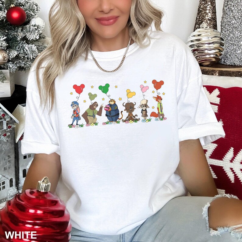 May include: White t-shirt featuring a colorful cartoon design of several animal characters holding heart-shaped balloons. The characters are standing on a patch of grass with flowers. The word "WHITE" is printed at the bottom of the image.