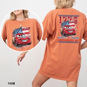 May include: An orange short-sleeve t-shirt featuring a graphic of a red race car with a United States flag background. The back design includes the text 1776 and UNITED STATES INDEPENDENCE DAY with a checkered border and red star accents.
