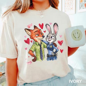 Może przedstawiać: Kremowa koszulka z akwarelową ilustracją Nicka Wilde'a i Judy Hopps z Zootopii, otoczona r&oacute;żowymi i czerwonymi sercami. Na dole koszulki wydrukowano słowo "IVORY".