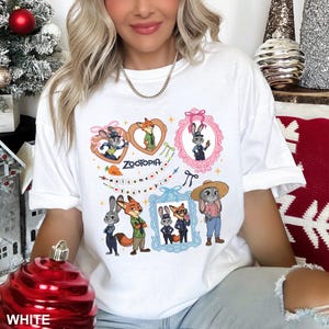 Op de afbeelding: Wit T-shirt met een kleurrijk cartoonontwerp met personages uit de film Zootopia. Het ontwerp bevat ingelijste afbeeldingen van de personages, het woord "Zootopia" en de zin "welcome to zootopia".
