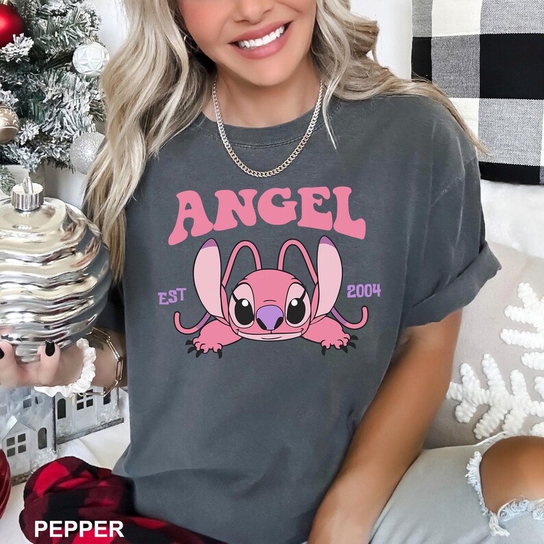 K&ouml;nnte beinhalten: Dunkelgraues T-Shirt mit einem rosa Cartoon-Engel-Design und dem Wort "ANGEL" dar&uuml;ber. Der Text "EST 2004" ist ebenfalls auf dem T-Shirt aufgedruckt. Das T-Shirt ist ein l&auml;ssiger Stil.
