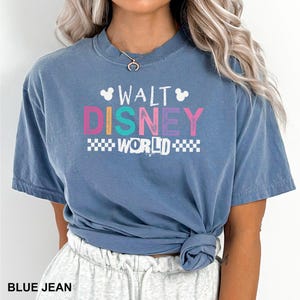 May include: A blue jean colored short-sleeve t-shirt featuring the text WALT DISNEY WORLD in a distressed white, pink, and teal font. The design includes two small white Mickey Mouse head silhouettes and a checkered pattern below the text.