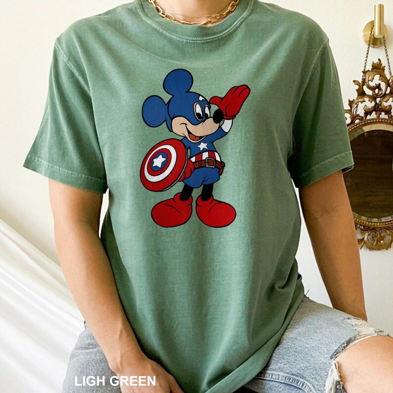 May include: A light green t-shirt featuring a cartoon Mickey Mouse dressed as Captain America. Mickey is wearing a blue costume with a white star, red boots, and holding a shield. The text "LIGH GREEN" is at the bottom of the image.