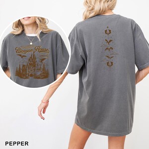 May include: Pepper-colored t-shirt featuring a brown graphic of a castle, trees, and flying creatures with the text "Rierson House". The back of the shirt displays a vertical design of stylized creatures and clouds.