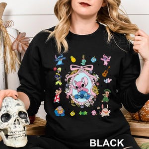 May include: Black crewneck sweatshirt featuring a colorful graphic of the character Stitch and other characters from the animated movie. The design includes a central oval frame with Stitch and Angel, surrounded by flowers and other elements. The word "BLACK" is printed at the bottom.