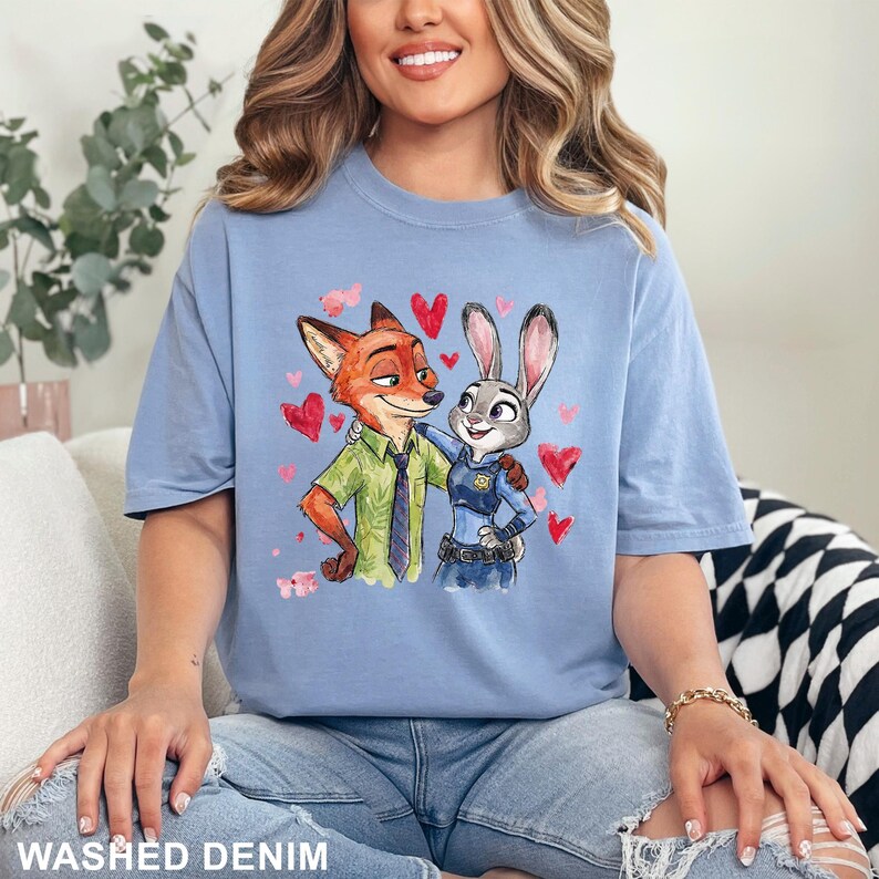 Może przedstawiać: Jasnoniebieska koszulka z akwarelową ilustracją Nicka Wilde'a i Judy Hopps z Zwierzogrodu, otoczona czerwonymi sercami. Na dole koszulki napis "WASHED DENIM".