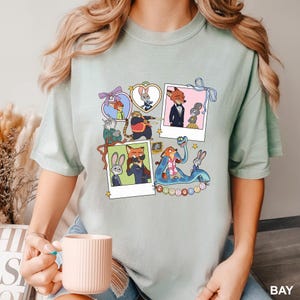 May include: Pale green t-shirt with a collage of cartoon characters in polaroid frames. The characters include a fox, rabbit, and other animals. The word "ZOOTOPIA" is printed on the bottom right. The shirt is made of a soft, comfortable material.
