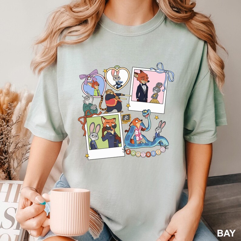 May include: Pale green t-shirt with a collage of cartoon characters in polaroid frames. The characters include a fox, rabbit, and other animals. The word "ZOOTOPIA" is printed on the bottom right. The shirt is made of a soft, comfortable material.