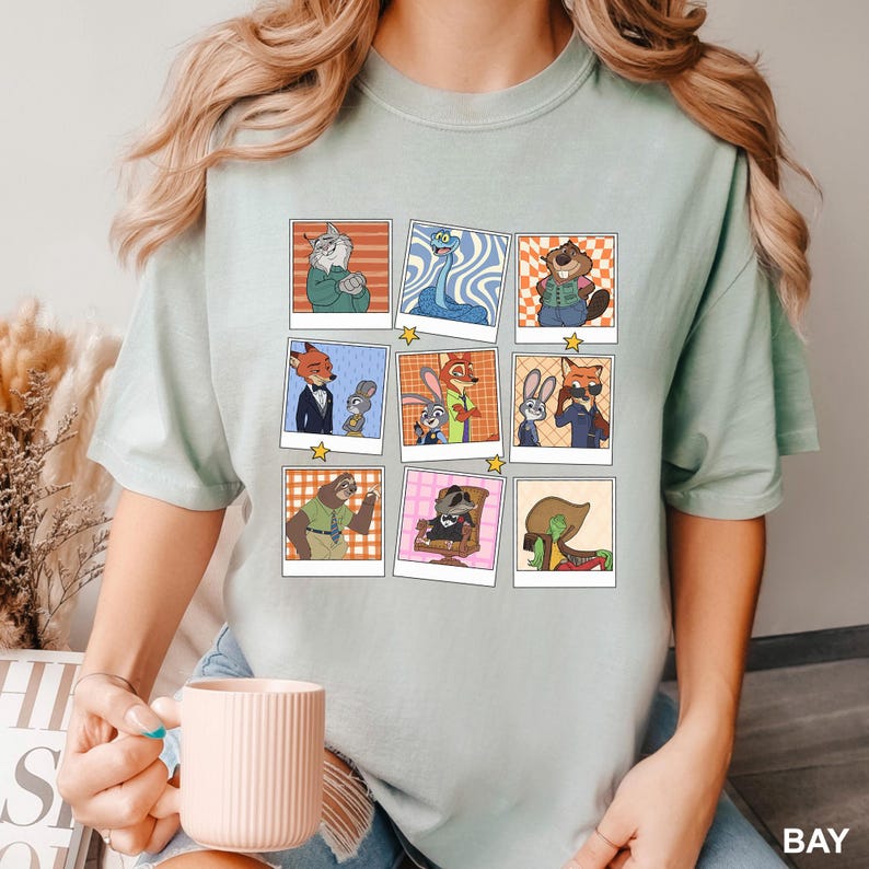 May include: Light green t-shirt featuring a grid of cartoon character portraits in polaroid-style frames. The characters include a fox in a suit, a sloth, and a raccoon. The word "BAY" is printed in the bottom right corner.