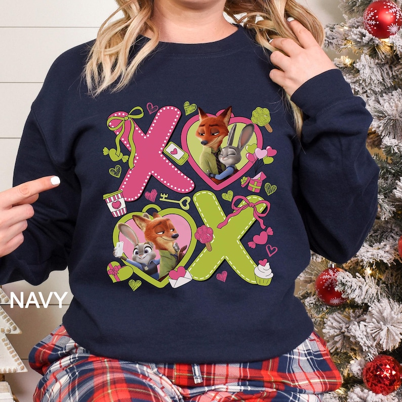 Puede incluir: Sudadera azul marino con un dise&ntilde;o gr&aacute;fico de los personajes Nick Wilde y Judy Hopps de Zootopia. El dise&ntilde;o incluye corazones rosas y verdes, las letras "X" y "O", y otros elementos tem&aacute;ticos de San Valent&iacute;n.