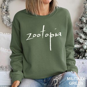 May include: An olive green sweatshirt with the word "zootopia" in white script across the chest. The sweatshirt has a crew neck. The text "MILITARY GREEN" is at the bottom right.