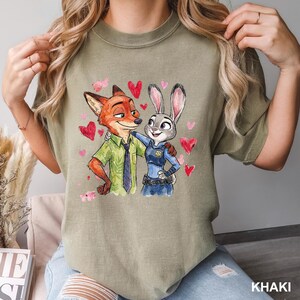 Może przedstawiać: Khaki koszulka z akwarelową ilustracją Nicka Wilde'a i Judy Hopps z Zootopii, otoczonych czerwonymi i r&oacute;żowymi sercami. Nick ma na sobie zieloną koszulę i krawat, a Judy mundur policjantki.