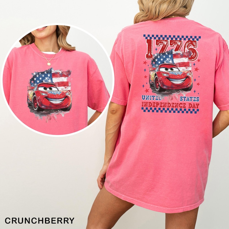 May include: A bright pink t-shirt featuring a graphic of a red race car with a smiling face, positioned in front of a stylised American flag. The back design includes the text 1776 and UNITED STATES INDEPENDENCE DAY with a checkered border.