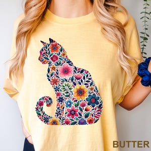 May include: A yellow t-shirt featuring a floral cat design. The cat silhouette is filled with colorful flowers in shades of pink, orange, blue, and yellow. The word "BUTTER" is printed at the bottom right corner of the shirt.