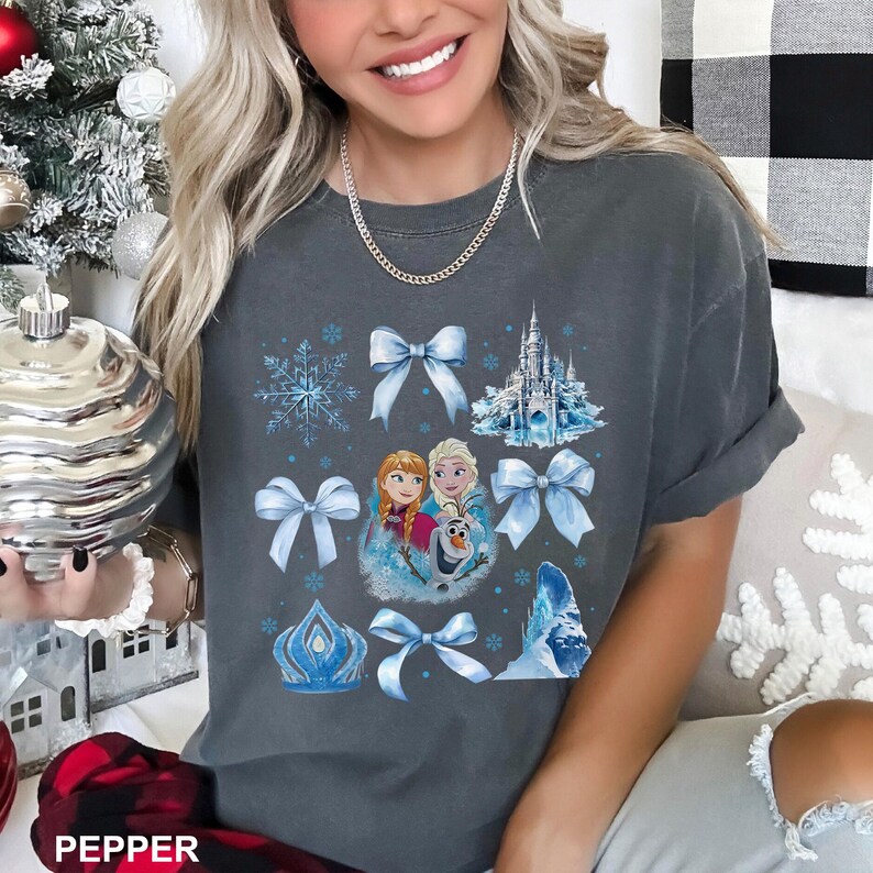 May include: Dark gray t-shirt featuring a graphic design with light blue bows, snowflakes, a castle, and characters. The word "PEPPER" is printed at the bottom of the image. The shirt is a comfortable, casual style.