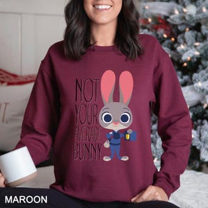 Op de afbeelding: Bordeaux sweatshirt met de tekst "NOT YOUR AVERAGE BUNNY" en een cartoon konijn karakter. Het konijn is grijs met roze oren, draagt een blauw uniform en houdt een aktetas vast. De sweatshirt heeft een effen bordeauxrode kleur.