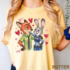 Może przedstawiać: Ż&oacute;łta koszulka z akwarelową ilustracją Nicka Wilde'a i Judy Hopps z Zootopii, otoczona czerwonymi i r&oacute;żowymi sercami. Słowo "BUTTER" jest wydrukowane w prawym dolnym rogu.
