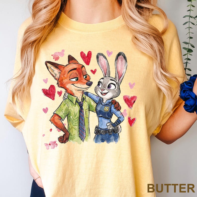 Może przedstawiać: Ż&oacute;łta koszulka z akwarelową ilustracją Nicka Wilde'a i Judy Hopps z Zootopii, otoczona czerwonymi i r&oacute;żowymi sercami. Słowo "BUTTER" jest wydrukowane w prawym dolnym rogu.