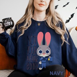 Op de afbeelding: Marineblauw sweatshirt met een cartoon konijn ontwerp en de tekst "NOT YOUR AVERAGE BUNNY". Het konijn heeft roze oren en houdt een blauwe tas vast. Het woord "NAVY" staat onderaan het sweatshirt.