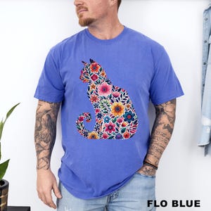 May include: A blue t-shirt featuring a floral cat design. The cat silhouette is filled with colorful flowers in shades of pink, yellow, blue, and red. The text "FLO BLUE" is printed at the bottom of the image.