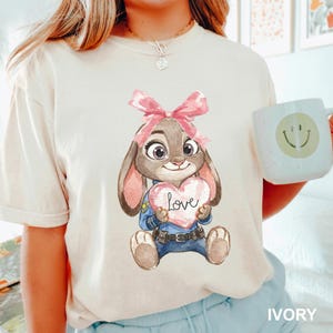 May include: Ivory-colored t-shirt featuring a watercolor illustration of a bunny holding a heart-shaped sign with the word "Love." The bunny wears a police uniform and a pink bow. A white mug with a smiley face is held in the hand.