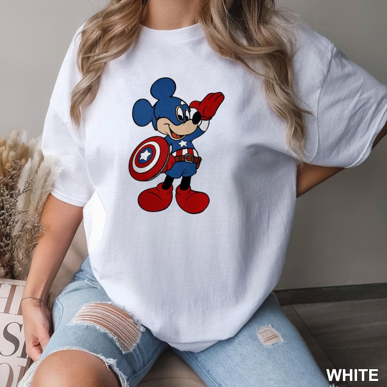 May include: White t-shirt featuring a cartoon Mickey Mouse dressed as Captain America. Mickey is in a blue and red costume, holding a shield. The shirt has a crew neck and short sleeves. The word "WHITE" is printed at the bottom right.