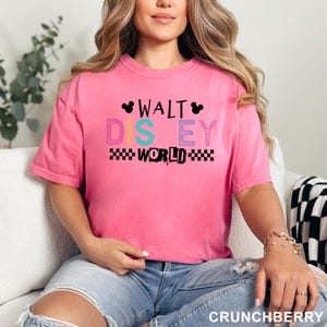 May include: A bright pink short-sleeve t-shirt featuring the text WALT DISNEY WORLD in a mix of black, teal, and purple colors. The design includes two small black Mickey Mouse ear silhouettes and a black and white checkered pattern border.
