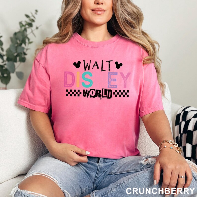 May include: A bright pink short-sleeve t-shirt featuring the text WALT DISNEY WORLD in a mix of black, teal, and purple colors. The design includes two small black Mickey Mouse ear silhouettes and a black and white checkered pattern border.