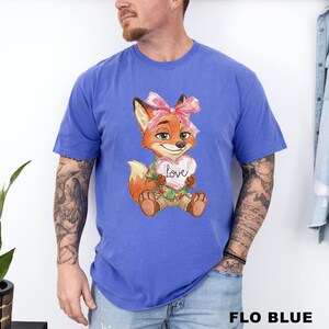 May include: Flo blue t-shirt featuring a cartoon fox holding a heart with the word "love" on it. The fox has a pink bow and is wearing a Hawaiian shirt. The text "FLO BLUE" is at the bottom of the image.