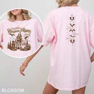 May include: Light pink t-shirt with a brown graphic on the front featuring a castle, dragons, and the text "Riorrson House". The back of the shirt has a vertical design of dragons and other symbols. The word "Blossom" is at the bottom.
