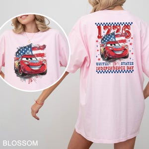 May include: A light pink t-shirt featuring a graphic of a red race car with a smiling face, positioned in front of a stylised American flag. The back design includes the text 1776 and UNITED STATES INDEPENDENCE DAY with red stars.