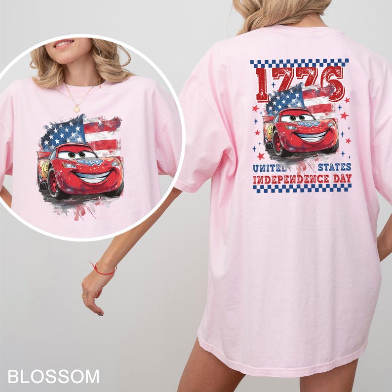 May include: A light pink t-shirt featuring a graphic of a red race car with a smiling face, positioned in front of a stylised American flag. The back design includes the text 1776 and UNITED STATES INDEPENDENCE DAY with red stars.