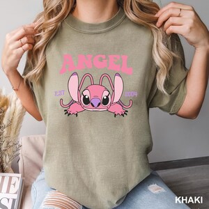 K&ouml;nnte beinhalten: Khaki-farbenes T-Shirt mit einer rosa Cartoonfigur und dem Wort "ANGEL" dar&uuml;ber. Die Figur hat gro&szlig;e Ohren und liegt. Der Text "EST 2004" befindet sich ebenfalls auf dem Shirt.