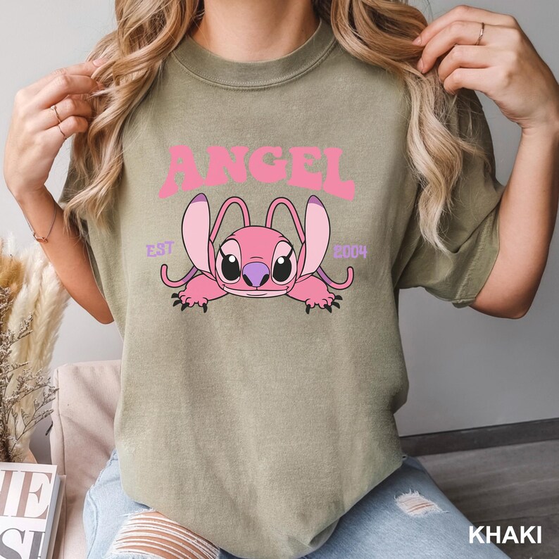 K&ouml;nnte beinhalten: Khaki-farbenes T-Shirt mit einer rosa Cartoonfigur und dem Wort "ANGEL" dar&uuml;ber. Die Figur hat gro&szlig;e Ohren und liegt. Der Text "EST 2004" befindet sich ebenfalls auf dem Shirt.