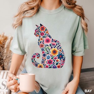 May include: A light green t-shirt featuring a floral cat silhouette. The cat is filled with colorful flowers in shades of pink, blue, yellow, and purple. The shirt has short sleeves and a crew neck.