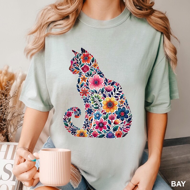 May include: A light green t-shirt featuring a floral cat silhouette. The cat is filled with colorful flowers in shades of pink, blue, yellow, and purple. The shirt has short sleeves and a crew neck.