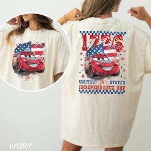 May include: An ivory t-shirt featuring a graphic of a red race car in front of an American flag. The text reads 1776, United States, and Independence Day, accompanied by red stars and a blue and white checkered border design on the back.