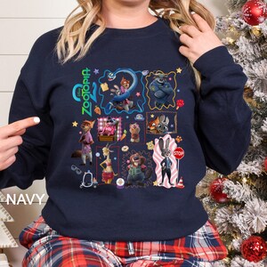 May include: Navy blue sweatshirt with a colourful collage of characters from the animated film "Zootopia." The design includes various characters in square and rectangular frames, with the word "Zootopia" in green. The word "NAVY" is printed on the bottom left.